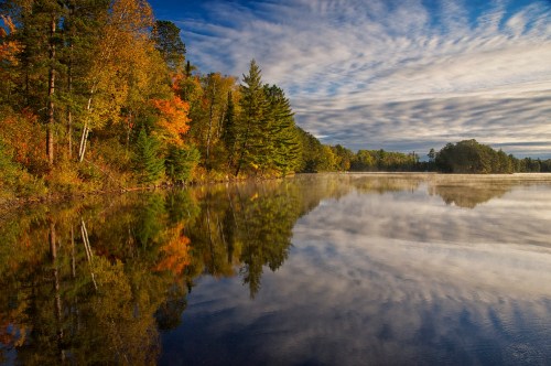A serene lake reflects colorful autumn trees and a cloudy sky, creating a peaceful natural landscape.