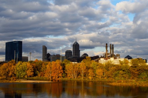 City skyline with autumn trees and a river, under a cloudy sky.