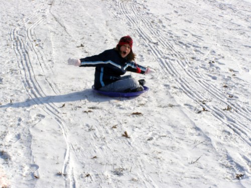 A child joyfully sledding down a snowy hill, wearing a winter coat and a red hat, with snow tracks in the background.