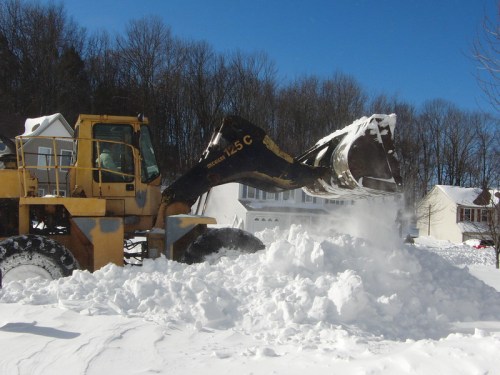 A snowplow clears a large pile of snow in a residential area under a clear blue sky.