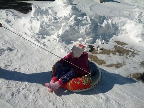 A child sits in a snow tube, being pulled on a snowy path, wearing a pink hat and colorful winter clothes.
