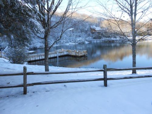 A serene winter scene with a snowy landscape, a calm lake, and a wooden dock surrounded by trees.