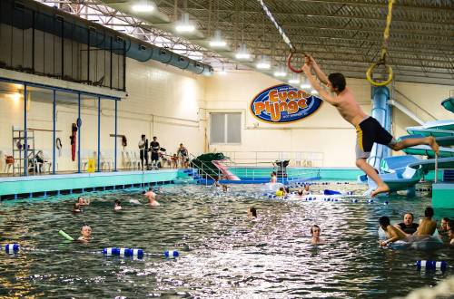 A boy swings from a rope above a pool, while others swim and play in the water at an indoor aquatic center.