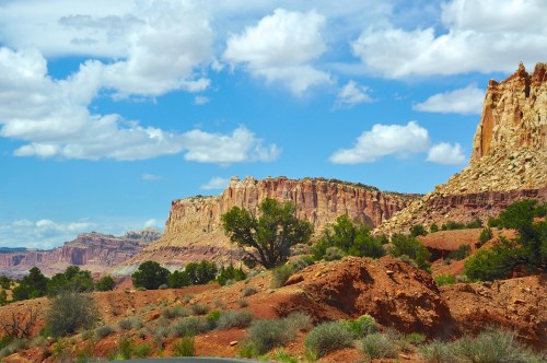 A scenic view of red rock cliffs under a blue sky with fluffy clouds, surrounded by desert vegetation.