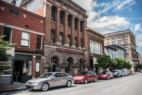 Historic buildings line a street with parked cars and trees, under a partly cloudy sky.