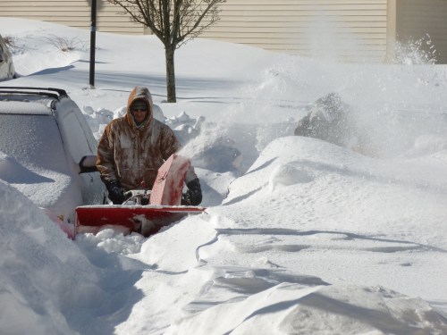 A person in a hooded jacket uses a snowblower to clear a snow-covered driveway, surrounded by deep snowdrifts.