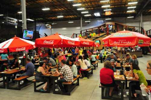 A busy indoor dining area with people eating at picnic tables under red umbrellas, surrounded by a lively atmosphere.