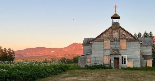 Abandoned church with a cross, surrounded by overgrown grass and mountains in the background during sunset.
