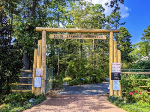 Entrance to South Arkansas Arboretum, featuring a wooden archway and signs, surrounded by lush greenery.