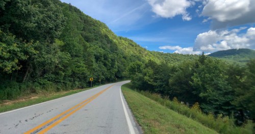 A winding road surrounded by lush green hills and a blue sky with fluffy clouds.