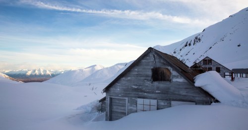 A weathered wooden cabin covered in snow, surrounded by mountains under a blue sky.