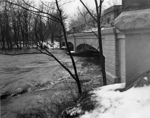 A river flows rapidly under a stone bridge, surrounded by bare trees and snow-covered ground.