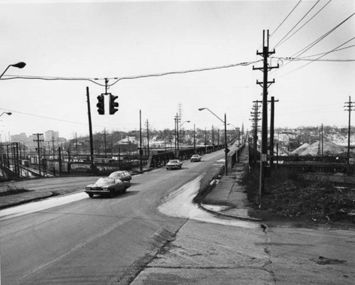 Black and white photo of a street intersection with traffic lights, power lines, and distant buildings.