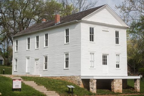 A historic white house on stone pillars, surrounded by greenery and a clear sky. Steps lead up to the entrance.
