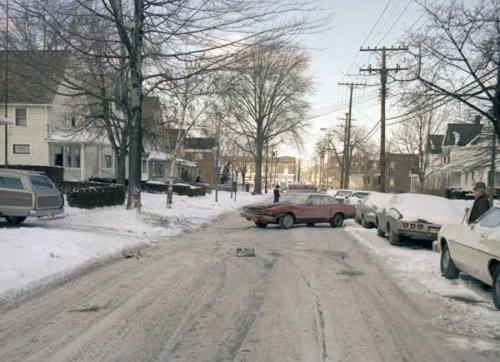 A snowy street scene with parked cars and a red car partially blocking the road, surrounded by bare trees and houses.