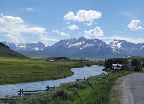 A scenic view of a river winding through green fields, with snow-capped mountains and blue skies in the background.