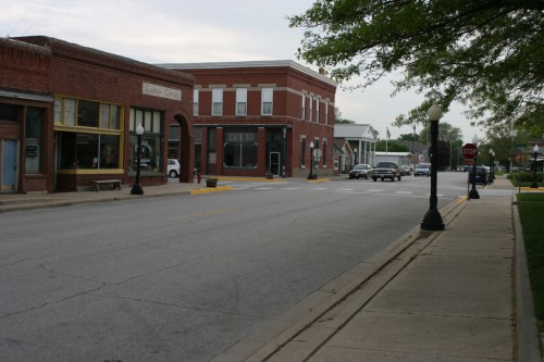 A quiet street scene featuring brick buildings, a stop sign, and trees lining the sidewalk on a cloudy day.