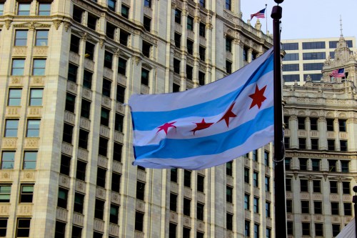 Chicago flag waving in front of historic buildings, featuring blue stripes and red stars against a clear sky.