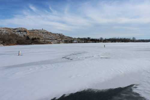 A frozen lake with a smooth, icy surface and distant hills under a blue sky with scattered clouds.