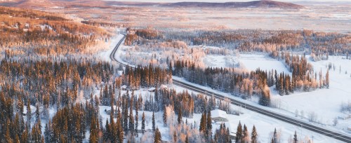 Aerial view of a snowy landscape with winding roads, dense forests, and distant mountains under soft sunlight.