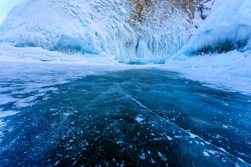A frozen lake with a smooth, dark surface, surrounded by icy cliffs and blue ice formations.