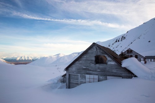 A weathered wooden cabin partially buried in snow, surrounded by mountains under a clear blue sky.