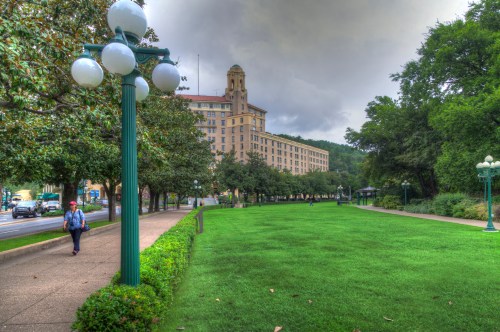 A park with green grass and trees, featuring a walking path and a historic building in the background under a cloudy sky.