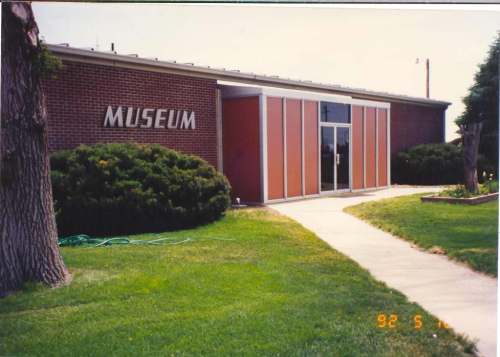 Exterior view of a museum building with a sign, surrounded by green grass and shrubs, on a sunny day.