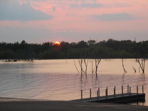 Sunset over a calm lake, with silhouetted trees and a dock extending into the water, creating a serene atmosphere.