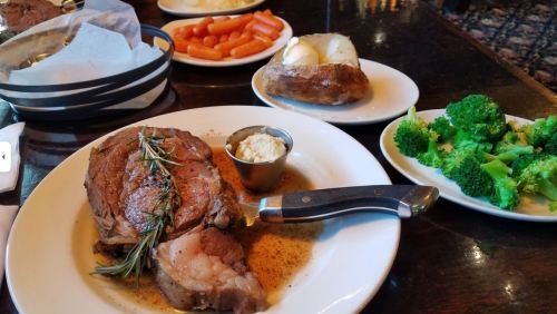 A plate with prime rib, rosemary, and butter, accompanied by baked potato, broccoli, and carrots on the side.