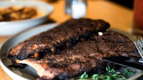 A close-up of grilled, charred ribs on a plate, garnished with fresh thyme, with a fork and knife beside them.