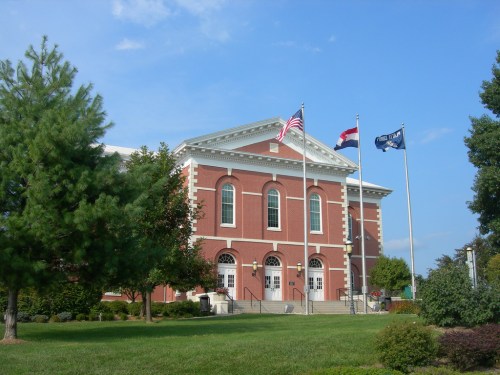 A brick building with large windows, flanked by flags, surrounded by green grass and trees under a blue sky.