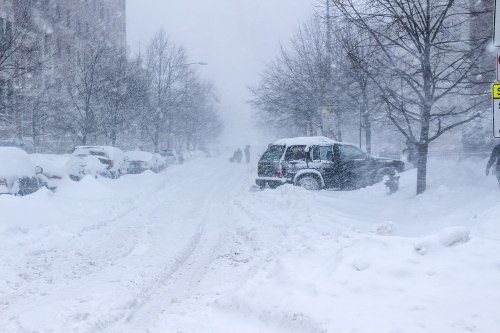 A snowy street during a blizzard, with cars buried in snow and people walking in the distance.