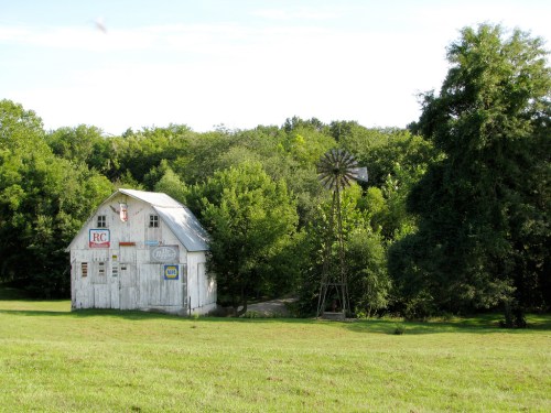 A rustic white barn with signs stands beside a windmill, surrounded by lush green trees and a grassy field.
