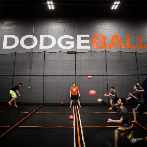 A group of people playing dodgeball on a trampoline court, with bright orange balls and a "DODGEBALL" sign on the wall.