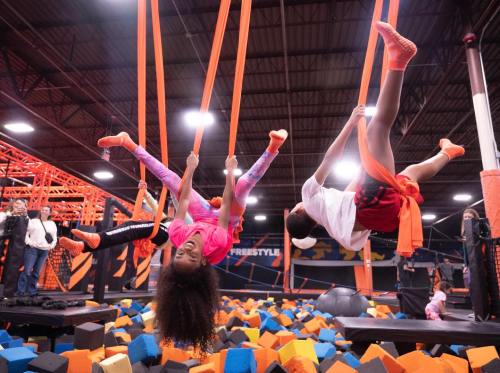 Children play on aerial silks above a foam pit in a vibrant indoor trampoline park.