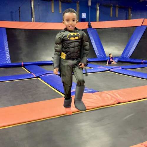 A young child dressed as Batman jumps on a trampoline in a colorful indoor trampoline park.