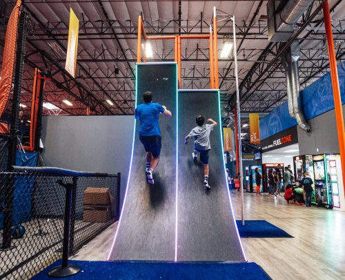 Two kids climbing a vertical wall in a trampoline park, with colorful lights and a lively atmosphere in the background.