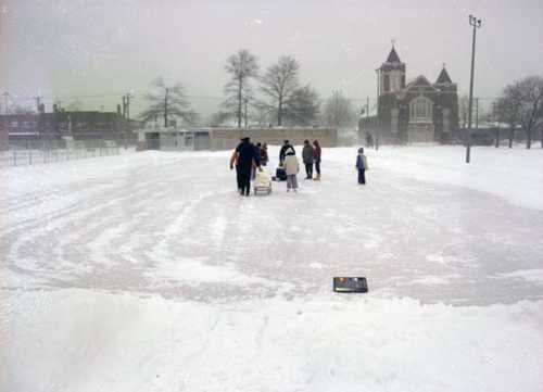 A snowy outdoor ice rink with people skating and walking, surrounded by trees and a church in the background.