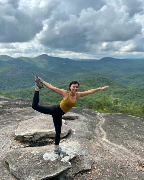 A person in a yellow top and black leggings balances on one leg on a rocky ledge, with mountains and clouds in the background.