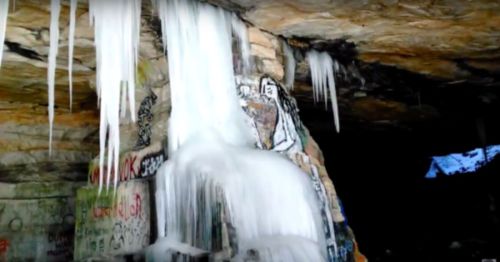 Icicles hang from a rocky cave ceiling, with colorful graffiti visible on the stone walls below.