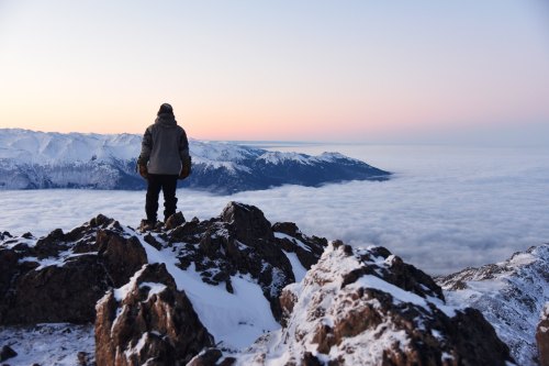 A person stands on a rocky mountain peak, overlooking a sea of clouds with snow-capped mountains in the background.
