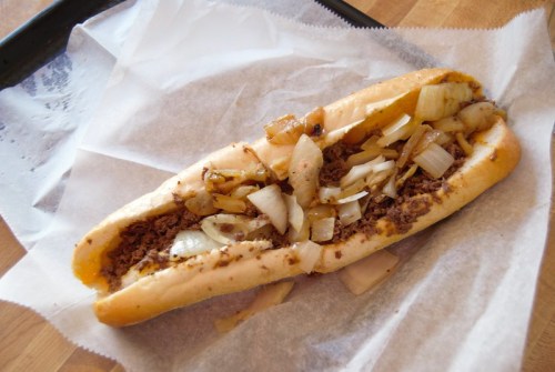 A close-up of a sandwich filled with seasoned meat and sautéed onions, served in a soft hoagie roll on parchment paper.