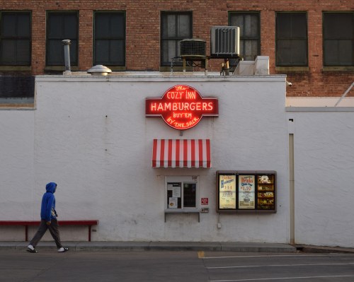 A person walks past the Cozy Inn, a hamburger restaurant with a bright neon sign and a striped awning.