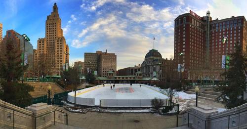 A panoramic view of an ice skating rink surrounded by buildings and trees under a cloudy sky.