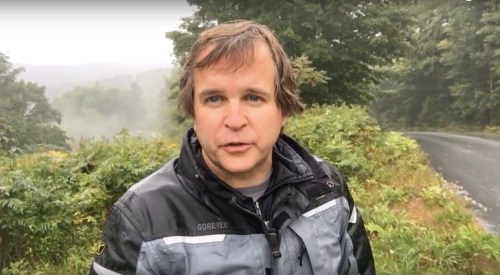 A man in a waterproof jacket stands outdoors, surrounded by greenery and misty hills in the background.