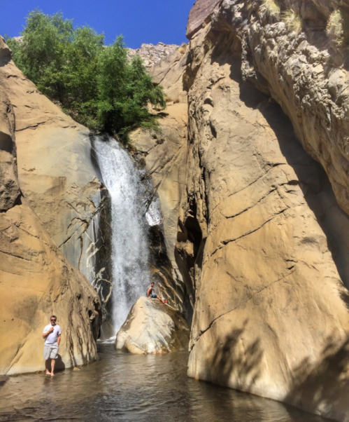 A waterfall cascades down rocky cliffs into a serene pool, with two people enjoying the natural scenery.