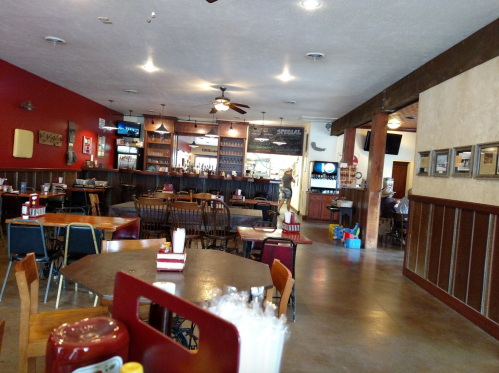 Interior of a casual restaurant with wooden tables, chairs, a bar area, and a few patrons in the background.