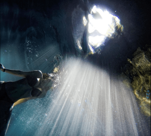 A person swims underwater, illuminated by beams of light filtering through the surface above.