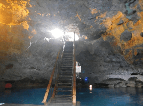 A wooden staircase leads up from a blue underground pool, illuminated by light filtering through a cave opening.